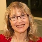 Christine is smiling and she has curly medium length light brown hair with blonde streaks. She is wearing red glasses and a red cardigan.