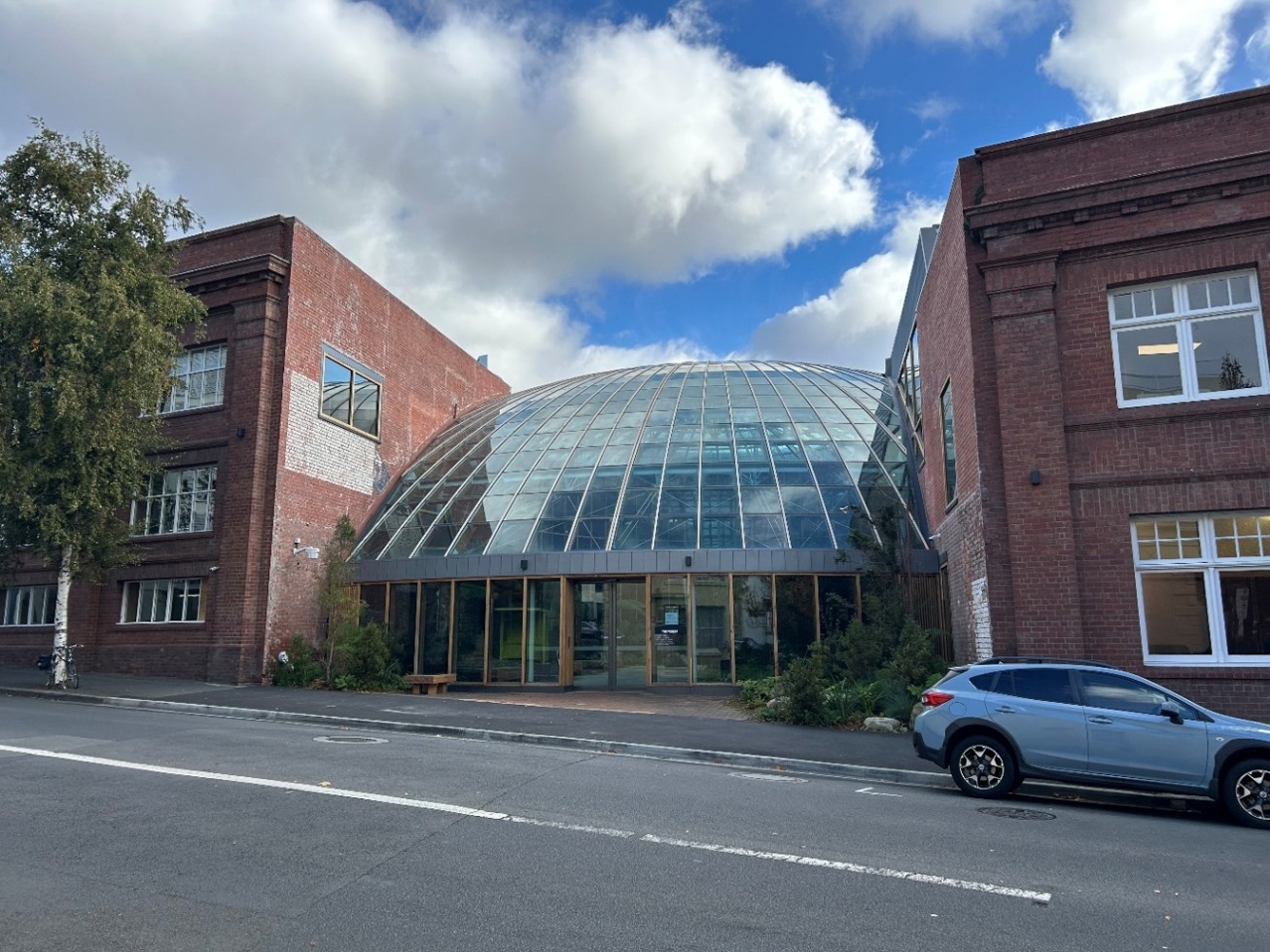 A building with three main sections, viewed from across the street. In the centre, a glass domed section, set back from the street; to each side, an old brick building with large white framed-windows.  