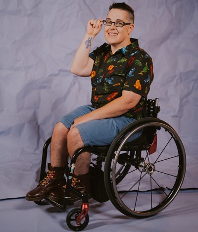 Jax is seated in their manual wheelchair smiling at the camera. They are wearing a black shirt with blue, green, orange and red frogs on it, blue shorts and maroon boots with rainbow laces. They have short black hair and are wearing glasses. Their right-hand is touching their glasses so you can see their wheelchair feminist tattoo. Image credit: Equality Institute and Eliza Allard photography