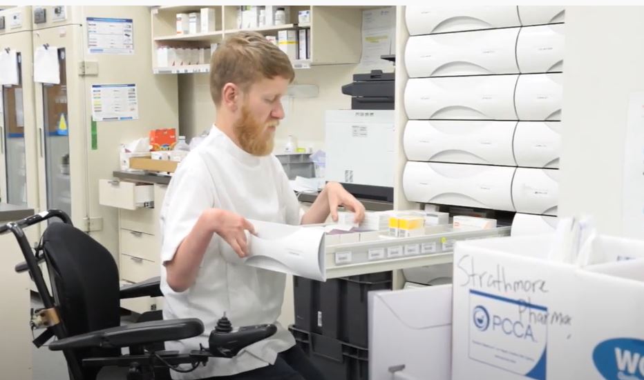 Sam flood at work in a pharmacy, picking up an item from an open drawer