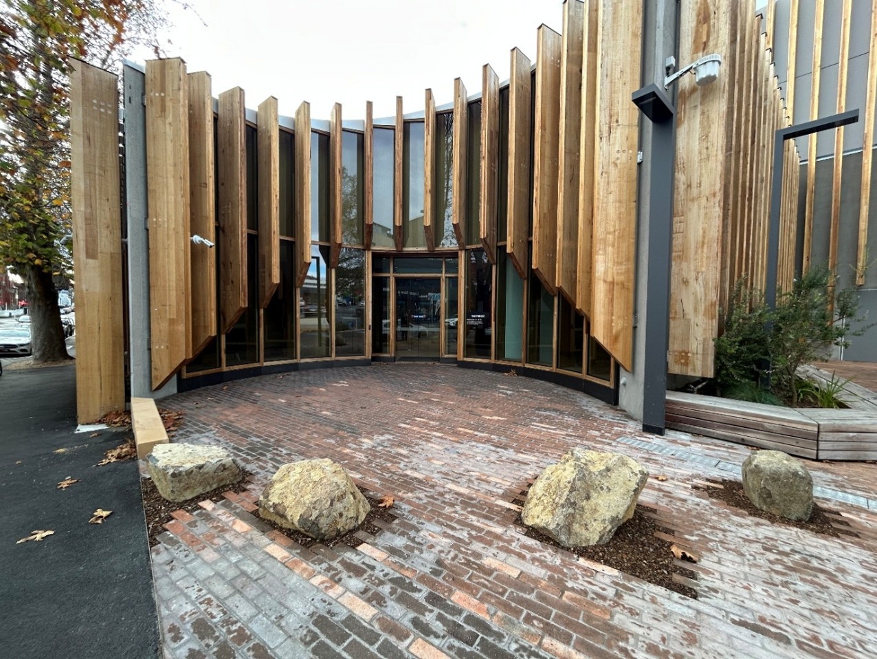A curved building decorated with a series of wooden panels facing inwards. The entrance area is paved with the same bricks as the parking area, creating a sense of continuity. The four boulders in the entrance zone are different shapes and sizes with uneven spacing between. The brick paving stops around each of the boulders and there are bark chips between the boulders and the bricks. 