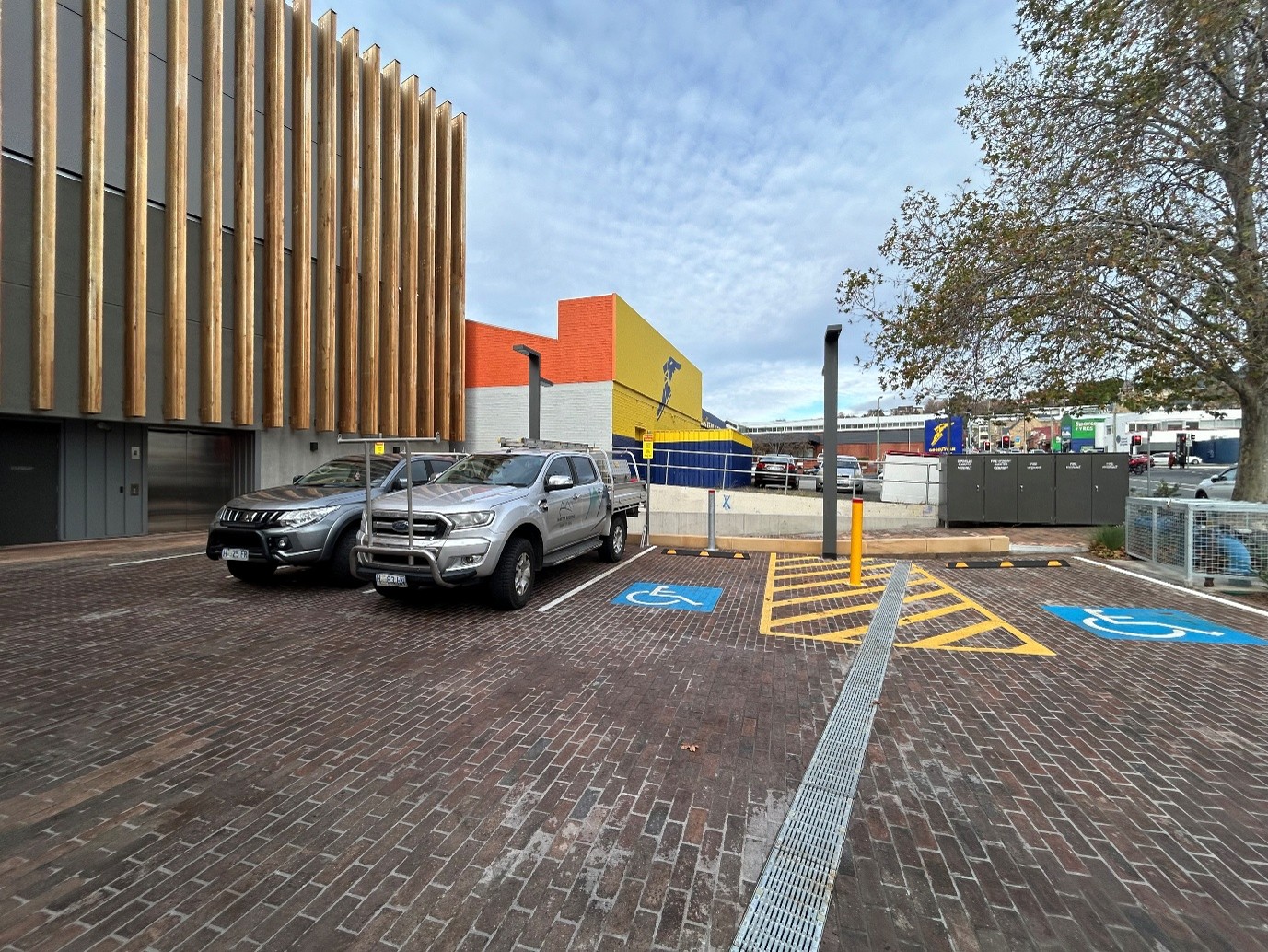 A wide open parking area with brick paving next to a wood panelled building. There are two disability parking spaces with a bollard and yellow lines on the ground between these parks, of equivalent space to another carpark. These parks are closest to the street. Beside these are three more parking spaces, closely aligned to each other. Two of these parks are marked as temporary disability parking. 