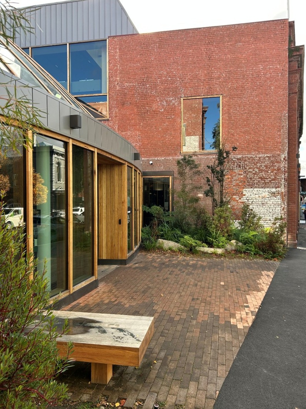A side view of the glass dome viewed from across the street earlier in the document. It’s set against a brick building, with paving and landscaping out the front. The front of the glass dome is made of curved glass, with a wooden inset indicating the entrance. 