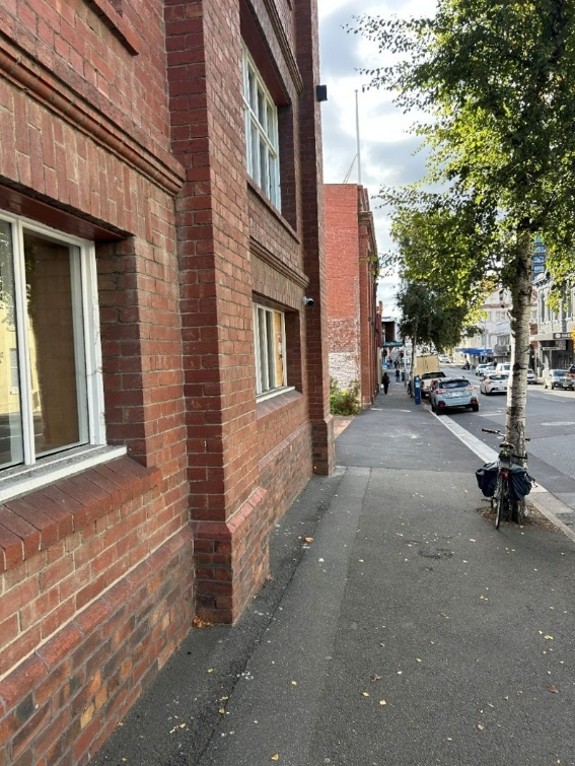 A few along a footpath, with a series of brick buildings on the left-hand side. There is a gap visible between the buildings, with some foliage poking out. 