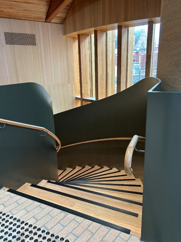 A set of spiral stairs curving to the left. There are tactile markers at the top, and wooden handrails on both sides of the staircase. 