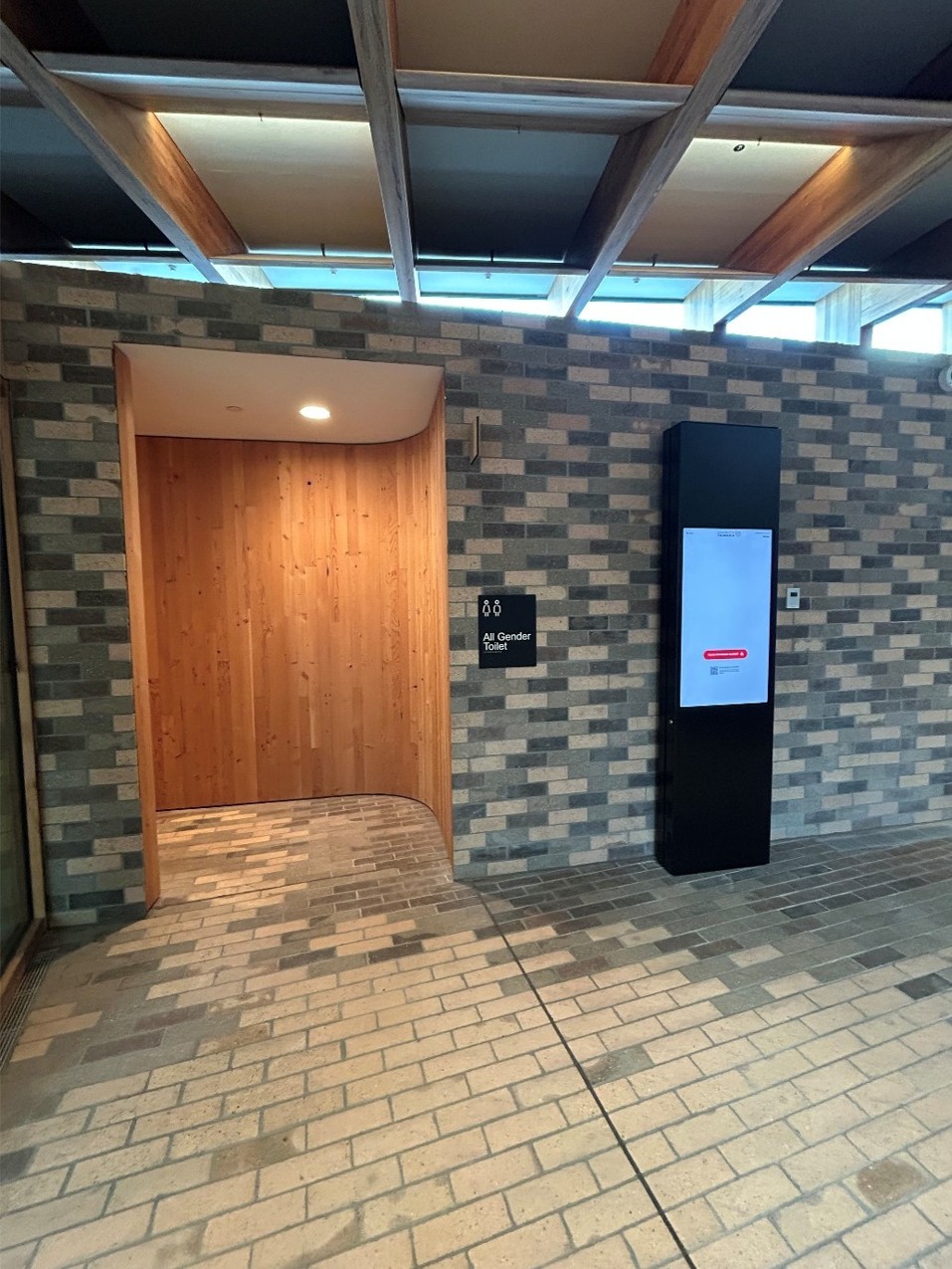 A wooden alcove set into a brick wall, leading off to the left into the bathrooms. On the brick wall outside the bathroom entry is a tall black panel with a large screen, displaying interactive maps. 
