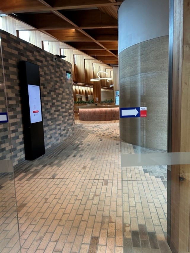 A view through glass sliding doors into a building area with muted colour tones. The left hand wall and the floor is all brick, and to the right is a large curved wall. Beyond, there is natural light, wooden panels, plants and some seating.