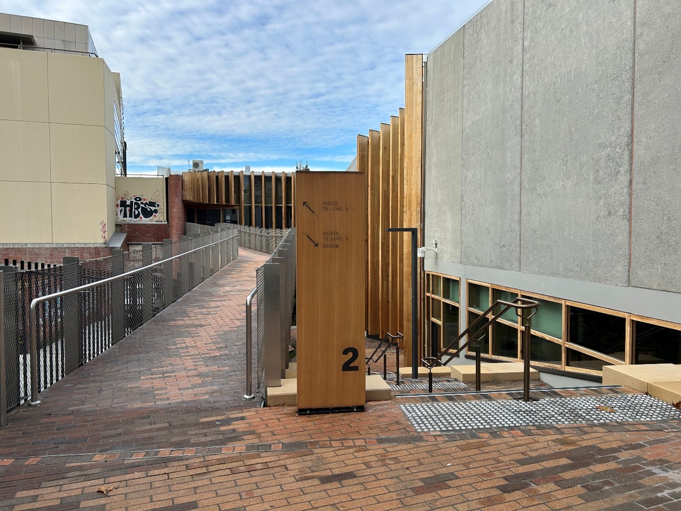 On the right-hand side of this photo is a spacious set of brick stairs leading downwards alongside a cement building facade. There are stair rails in the middle of each set of stairs and tactile markers at the top and bottom. To the left of the photo is a brick footbridge leading towards the upper rear entrance to the building. 