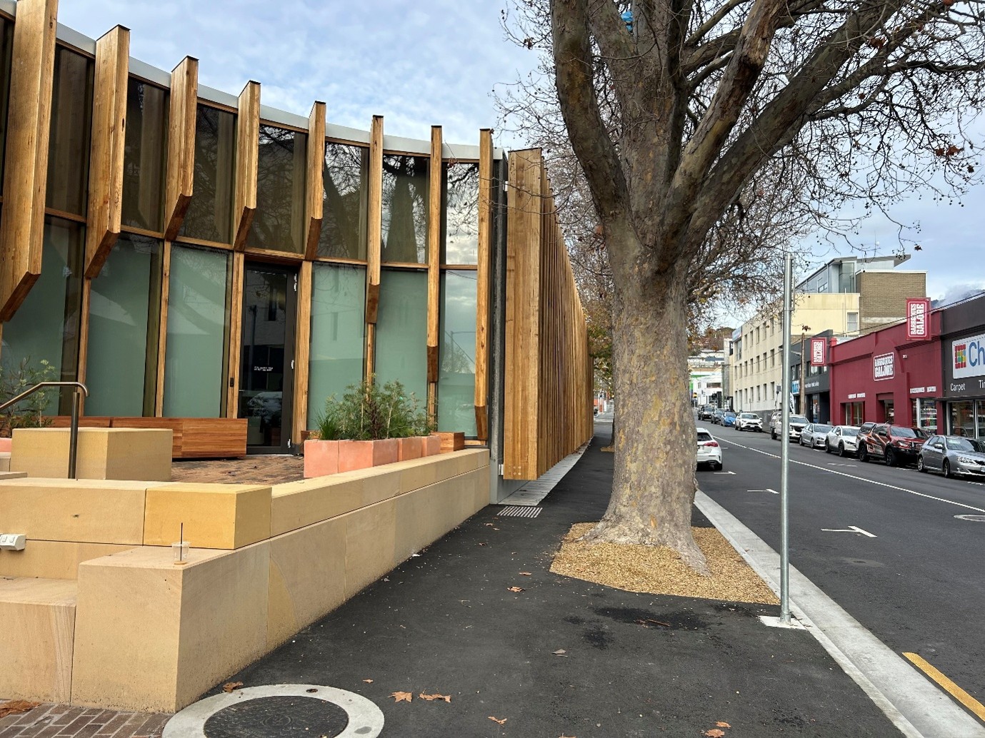 There are three main sections in this photo. To the right is Brisbane St with several parking spaces adjacent to the footpath. In the middle is the bitumen footpath leading along a narrow space between the building on the left and the large trees in the footpath. On the right is a building, decorated with large wooden panels, and in front of it a raised, brick-paved seating area with a sandstone retaining wall. 